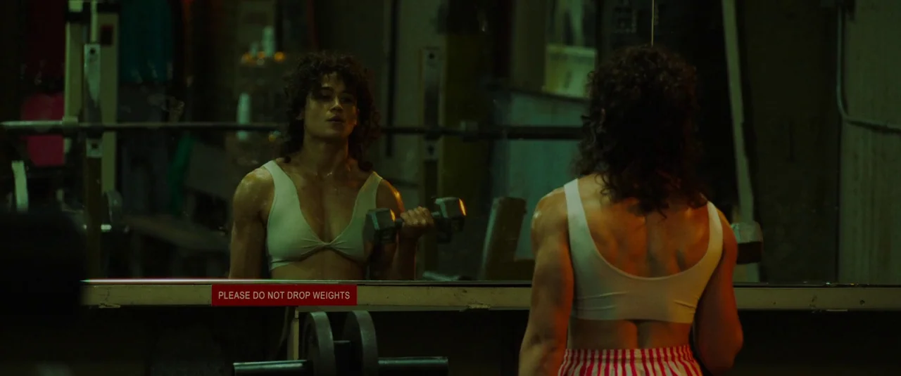 Love Lies Bleeding (2024) movie still: medium — A dimly lit room with workout equipment and a large mirror facing the subject. A muscular woman…
