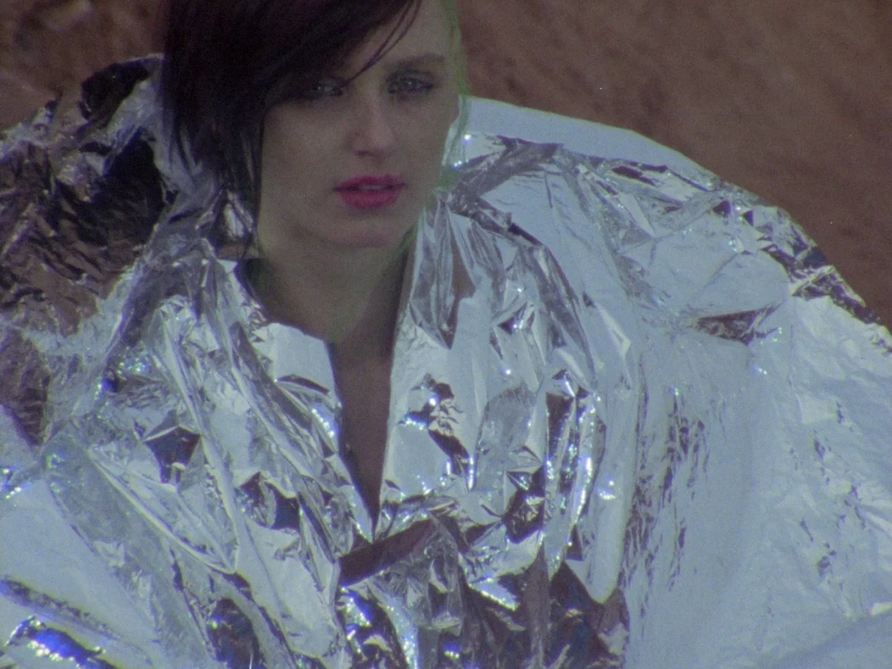 Decoder (1984) movie still: closeup — A person is wrapped in a shiny silver thermal blanket against a brown desert background. The subject is…