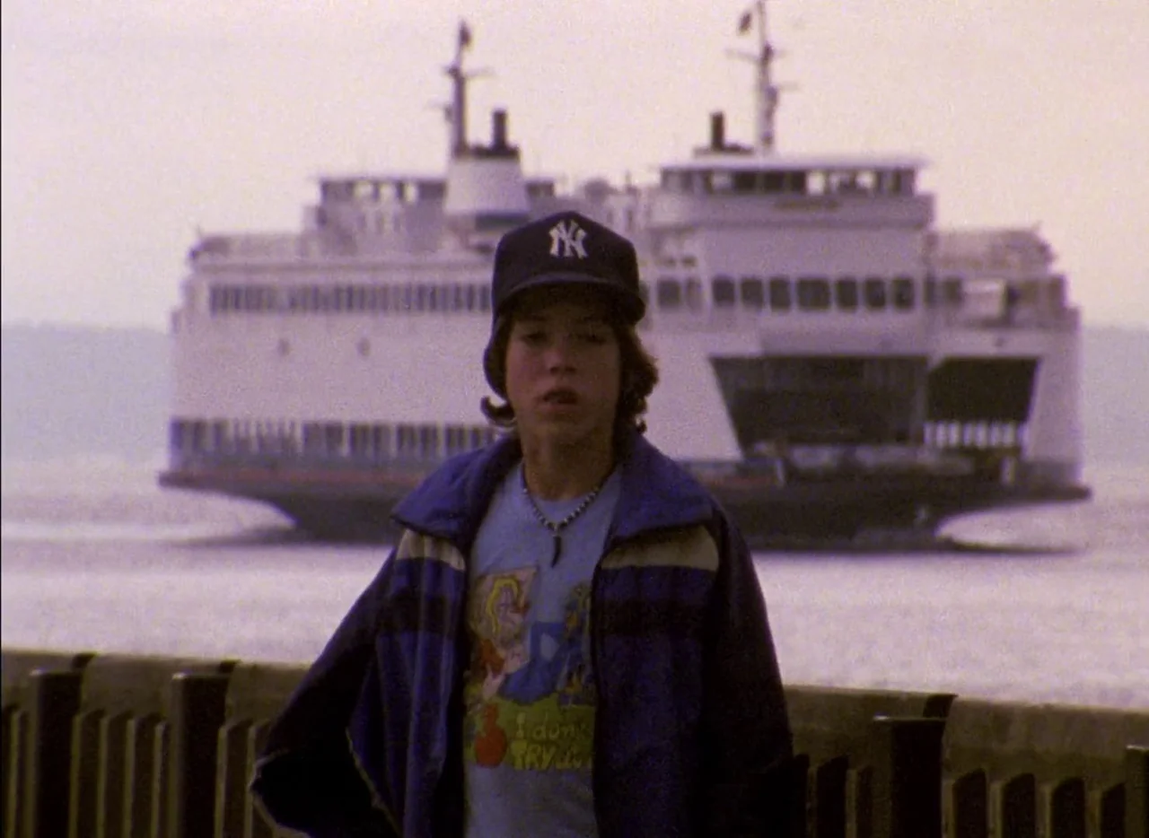 Streetwise (1984) movie still: medium — A teenager stands on a waterfront boardwalk with a large passenger ferry behind him. The water is calm and…
