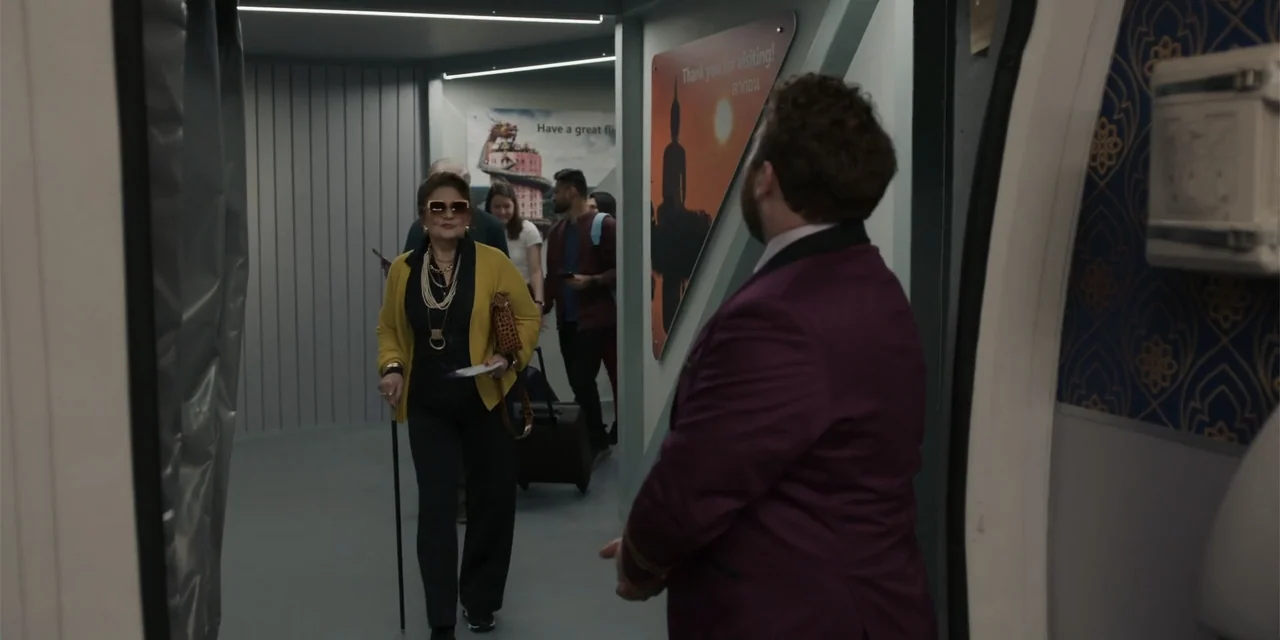 Fight or Flight (2025) movie still: fullbody — A modern airplane jet bridge features gray walls decorated with travel-themed wall decals. An elderly…
