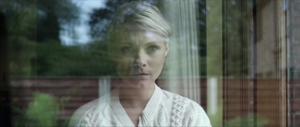 Kill List movie still: medium — A person is visible through a reflective glass pane looking out at a yard. The setting is a residential space with…