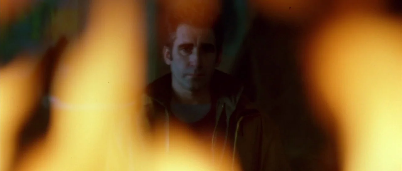 Isolation (2005) movie still: medium — A man stands in the dark illuminated by the orange glow of a fire. The out-of-focus foreground fire obscures…