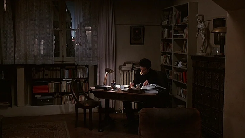 The Unbearable Lightness of Being (1988) movie still: wide — A dimly lit room filled with tall wooden shelves packed with numerous books. A man sits…