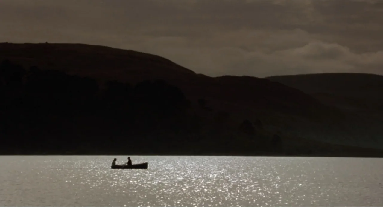 Dancing at Lughnasa (1998) movie still: wide — A vast lake stretches before dark rolling hills under an overcast sky. Two figures occupy a small…