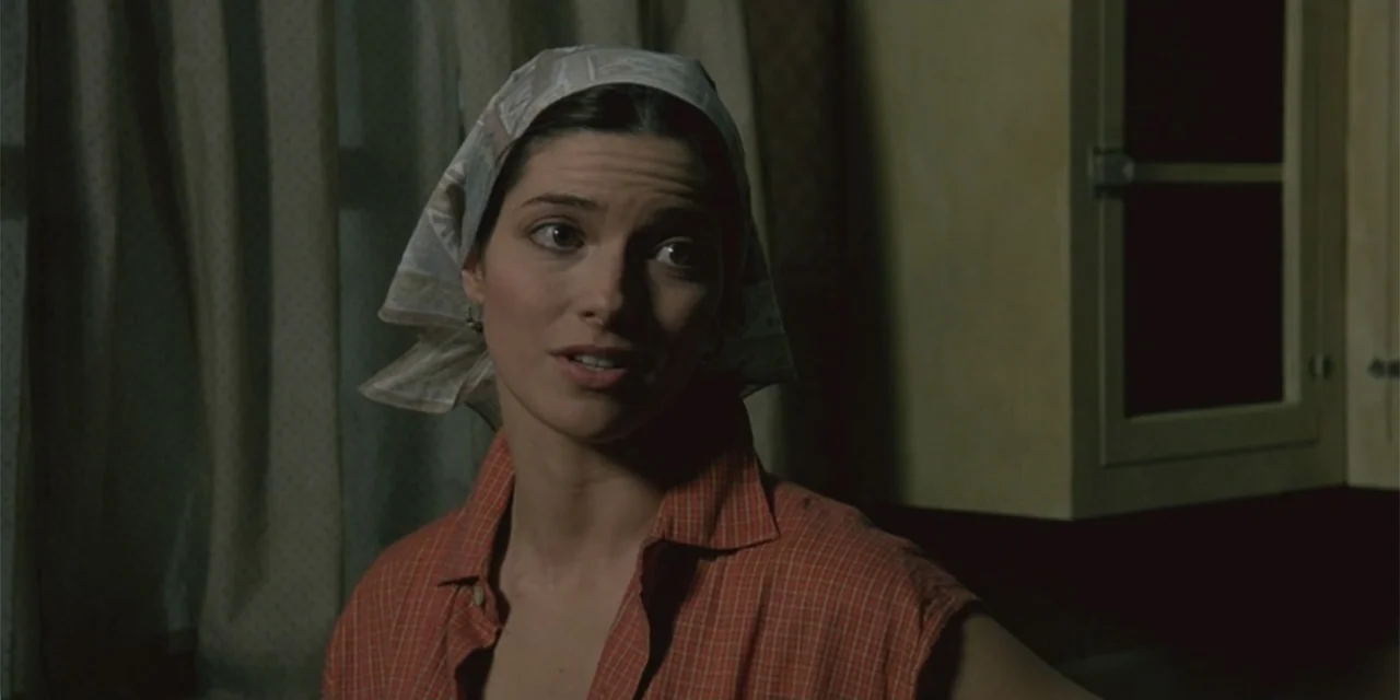 El sótano (2024) movie still: medium — A dimly lit room with simple cabinetry and a curtained window in the background. A woman wearing a patterned…