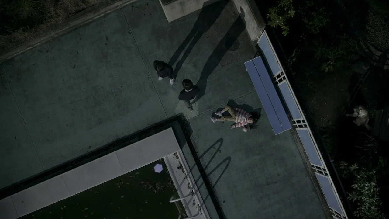 Confessions (2010) movie still: wide — A concrete outdoor terrace at night features a pool edge and a bench. Two figures stand while a small child…