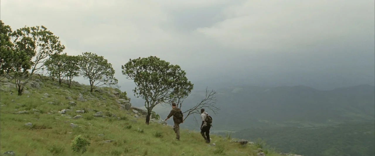 Blood Diamond (2006) movie still: wide — A grassy mountain slope features sparse trees and scattered rocks under an overcast sky. Two men with…
