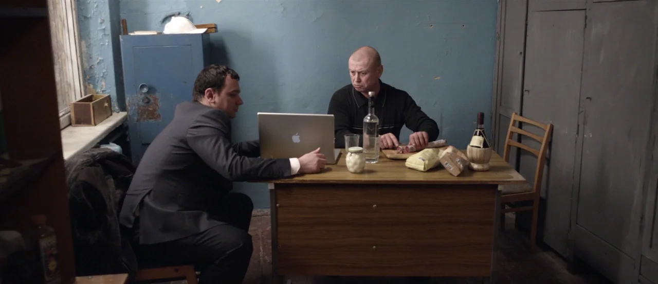 The Tribe (2014) movie still: medium — A spartan room has blue peeling paint on the walls and metal storage lockers. Two men sit at a small desk…