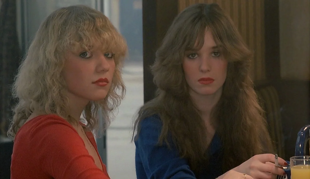 Angst (1983) movie still: medium — A diner booth with dark wood-paneled walls and a glass surface between the subjects. Two young women with…