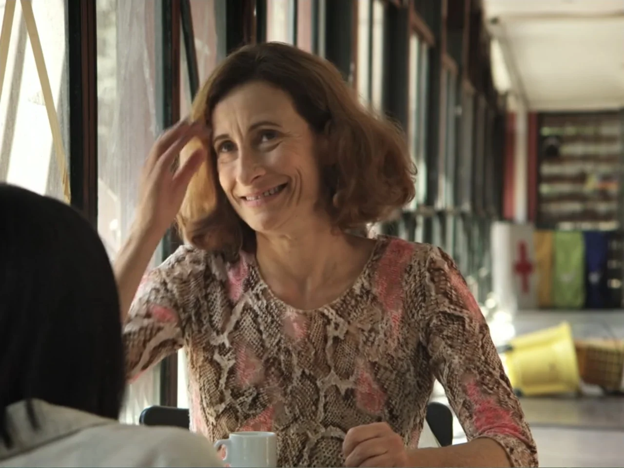 History and Geography (2024) movie still: medium — A woman sits at a table inside a diner booth near a large glass window. The woman reaches up with…