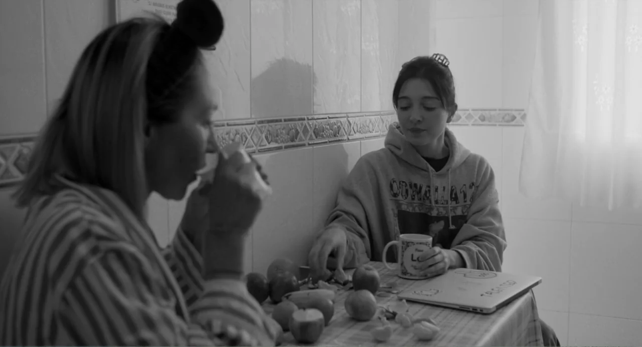 El Planeta (2021) movie still: medium — A tiled room with a table covered in scattered fruit and a laptop. Two young women sit across from each…