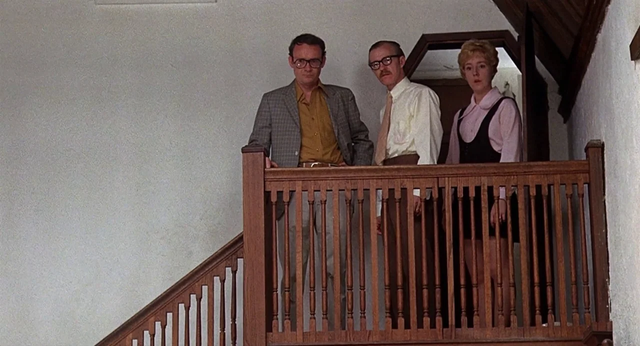 Taking Off (1971) movie still: medium — Three people stand together at the top of a wooden staircase inside a house. The landing features a simple…