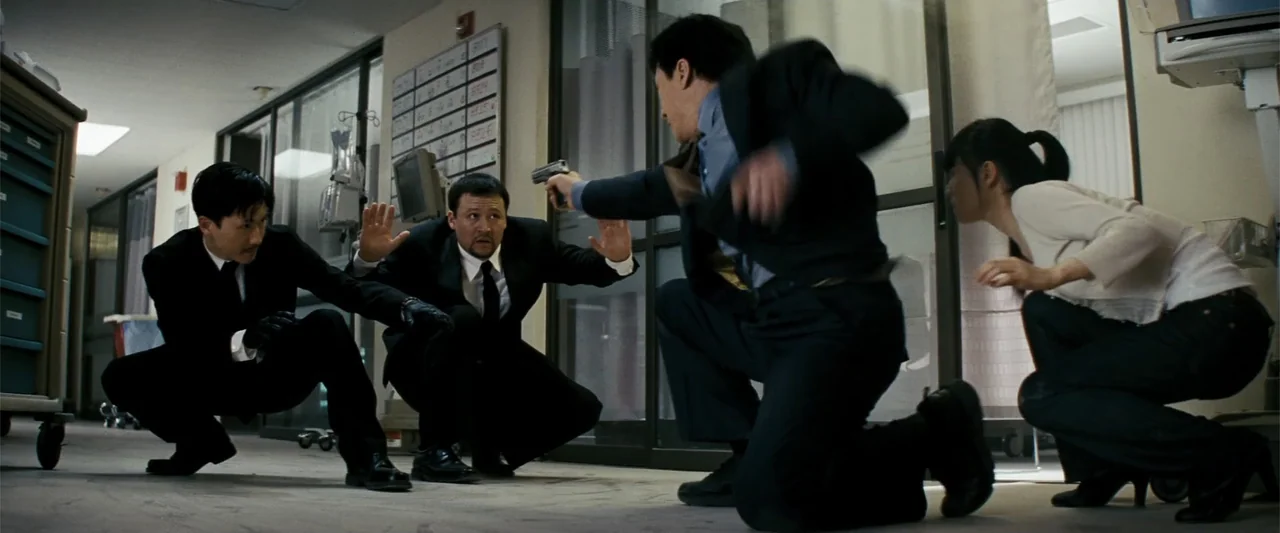 Rush Hour 3 (2007) movie still: medium — A sterile hospital hallway with glass doors and a wall-mounted patient chart board. Three men in suits…