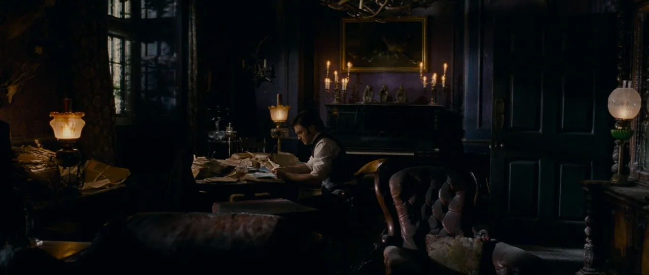 The Woman in Black (2012) movie still: wide — A dimly lit room with dark, ornate wallpaper and heavy furniture. A man sits at a desk cluttered with…