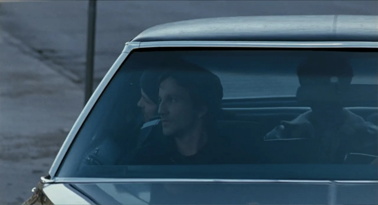 Colin Quinn: Red State, Blue State (2019) movie still: medium — A view through the windshield of a car parked on a paved street at dusk. A man in…