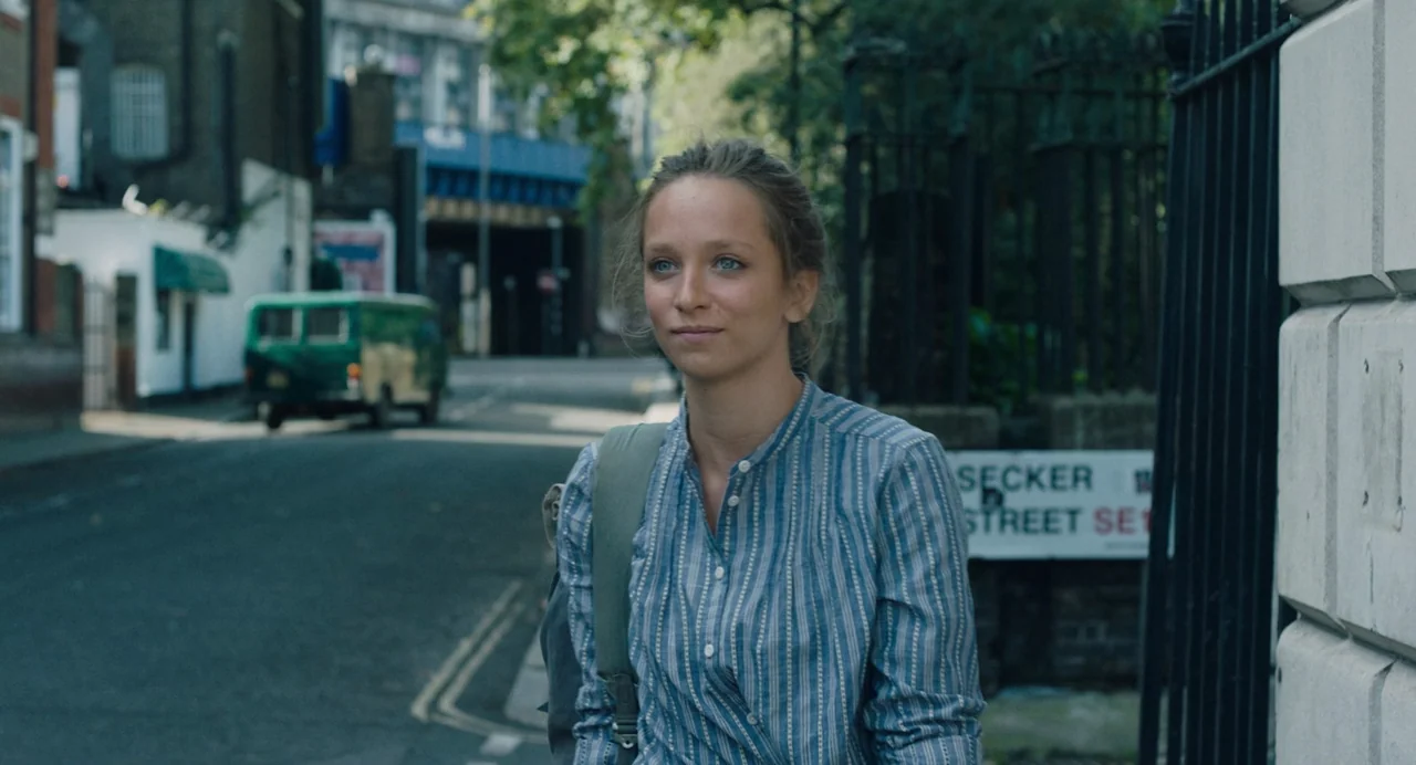 She Said (2022) movie still: medium — A quiet urban street in London features a green van in the distance. A woman in a striped shirt stands in the…