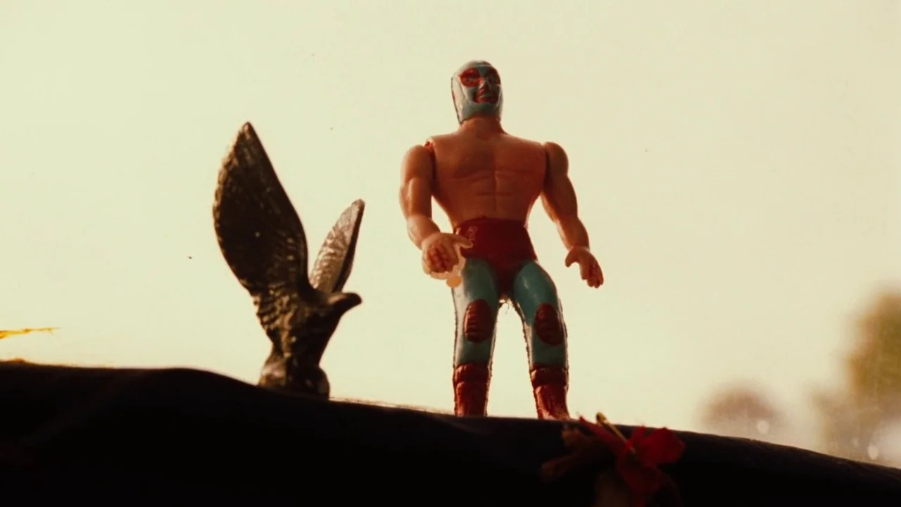 Nacho Libre (2006) movie still: closeup — A miniature plastic wrestler stands on the dark dashboard of a vehicle. A small toy eagle sits beside the…