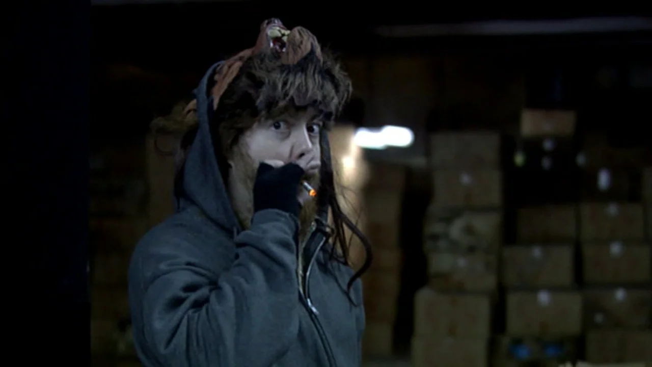 Murder Party movie still: medium — A dimly lit storage room filled with stacked cardboard boxes in the background. A man wearing an animal headpiece…