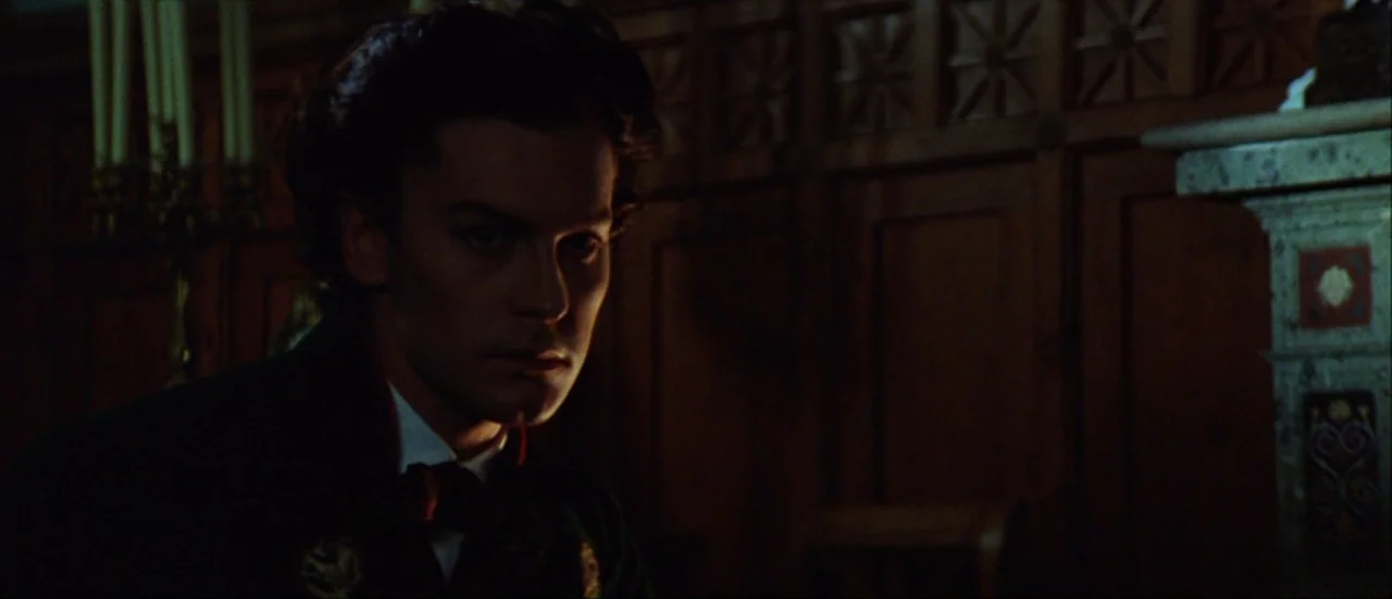 Ludwig (1973) movie still: closeup — A dimly lit room features wood-paneled walls and a fireplace in the background. A young man stands in the…