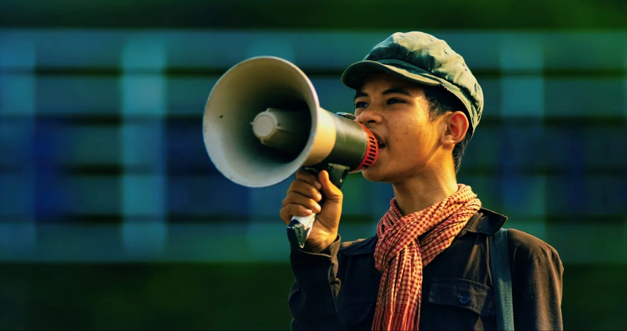 First They Killed My Father (2017) movie still: medium — A teenager stands outdoors against a blurred deep teal and green background. He holds a…