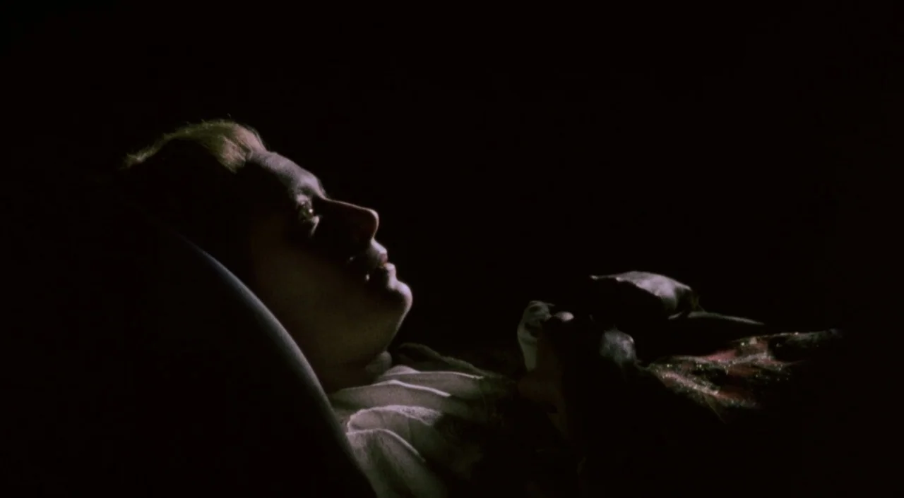 Orlando (1992) movie still: closeup — A person lies on a bed in a darkened room with black surroundings. The person is wearing a white nightgown and…