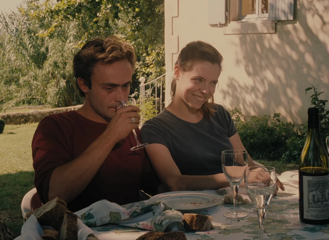 Autumn Tale (1998) movie still: medium — A wooden table set outdoors in front of a light-colored building with foliage behind. A young man and woman…