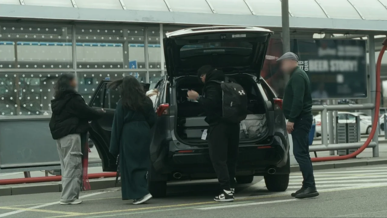 Bloody Trophy (2025) movie still: fullbody — An airport or transit pick-up zone features a dark car with its hatch open. A small group of people…