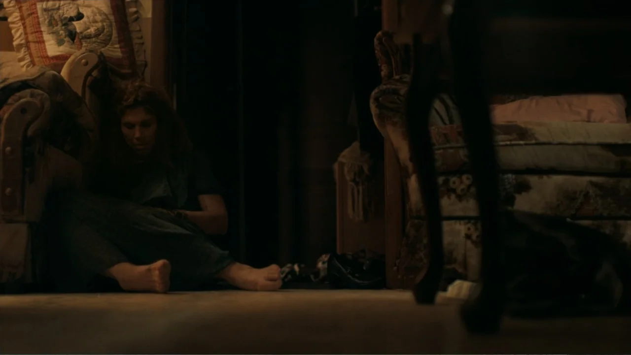 Alabama Snake (2020) movie still: medium — A dark living room contains a woman sitting on the carpet between two armchairs. She rests on the floor…