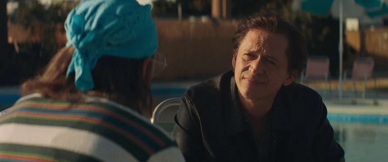 Honey Boy (2019) movie still: medium — A man in a black jacket sits poolside at sunset. He is looking across the table at another man wearing a blue…