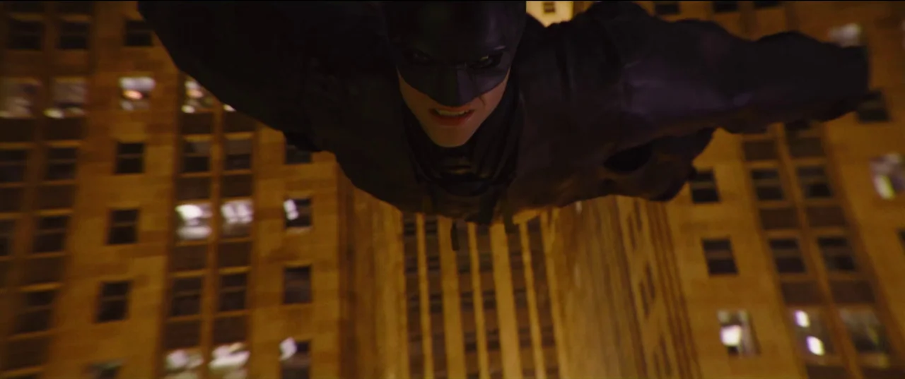The Batman (2022) movie still: closeup — A night view of a tall, dark skyscraper illuminated by glowing windows from below. A masked vigilante…