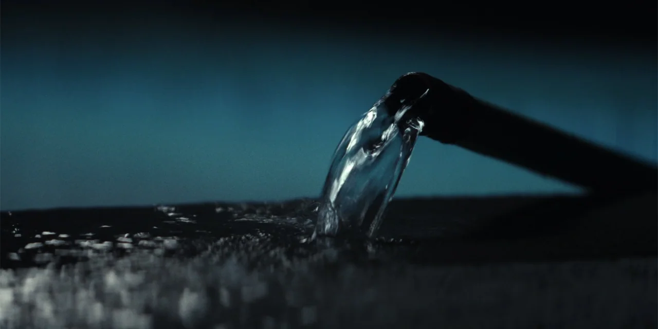 The Monkey (2025) movie still: closeup — A dark night setting features a pipe pouring water onto a dark surface. The clear liquid creates ripples…