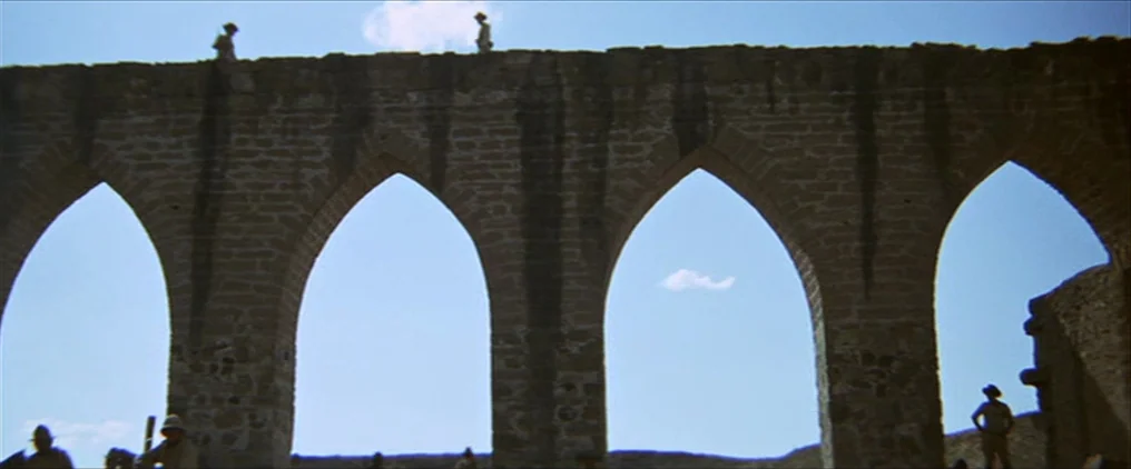 The Wild Bunch (1969) movie still: wide — An ancient stone aqueduct with several pointed arches stands against a clear blue sky. Several men wearing…