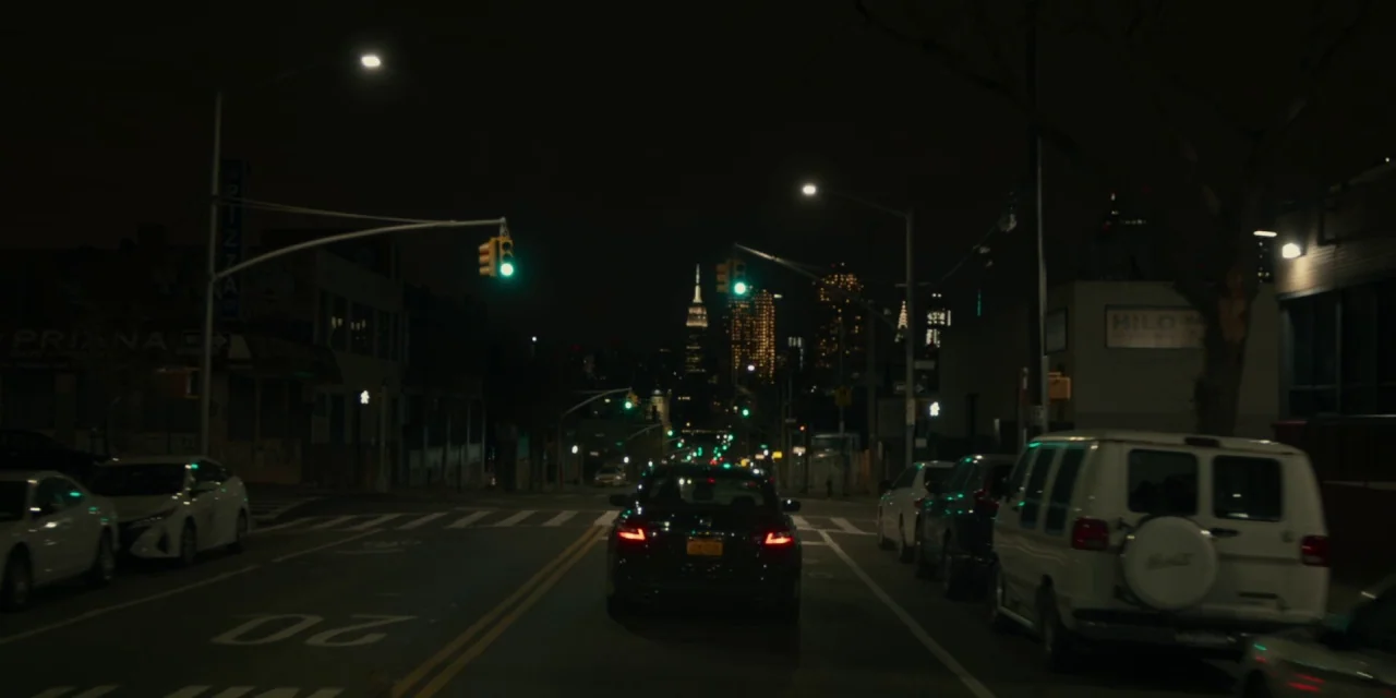 The Assistant (2019) movie still: wide — A quiet city street at night features rows of parked cars on both sides. In the distance, the illuminated…