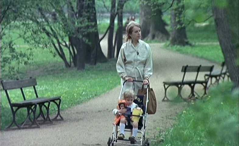 Blind Chance (1987) movie still: fullbody — A gravel path runs through a wooded area with mature trees and wooden benches. A woman walks along the…