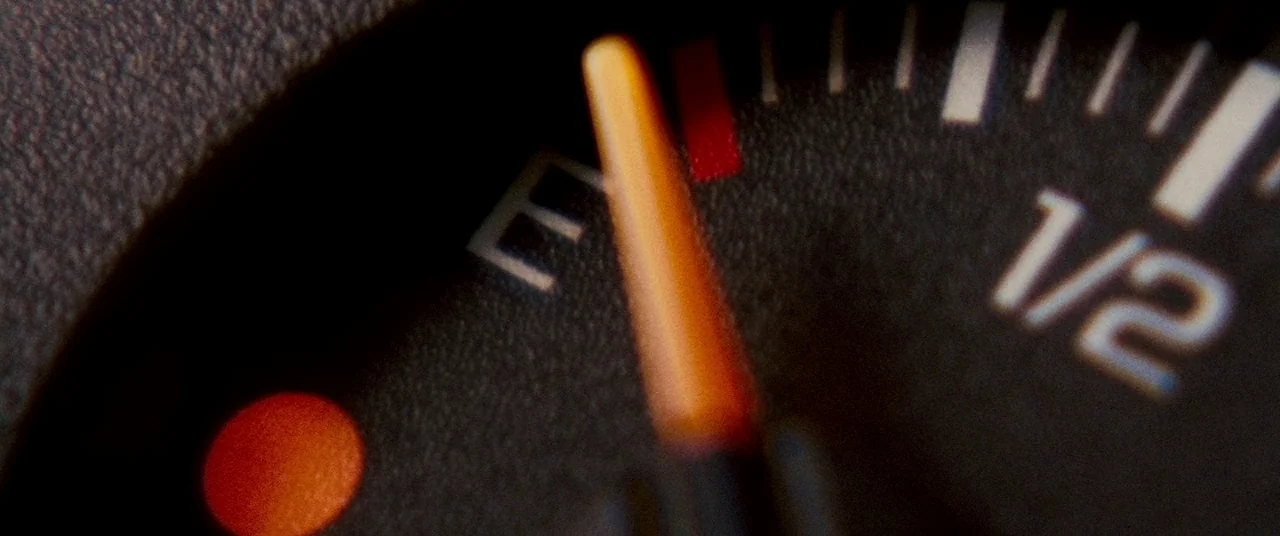 Carriers (2009) movie still: closeup — A dark, textured fuel gauge from a vehicle dashboard shows the needle on empty. A small circular red low-fuel…