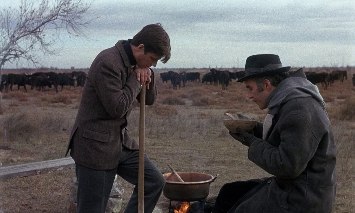 Belle De Jour (1967) movie still: fullbody — An open cattle pasture on a gray, overcast day contains a small cooking fire. Two men rest together…