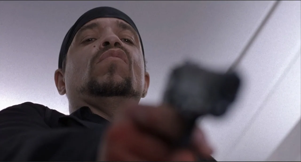 Air Rage (2001) movie still: closeup — An interior space with a plain white ceiling visible behind the subject. A man in a black skullcap aims a…