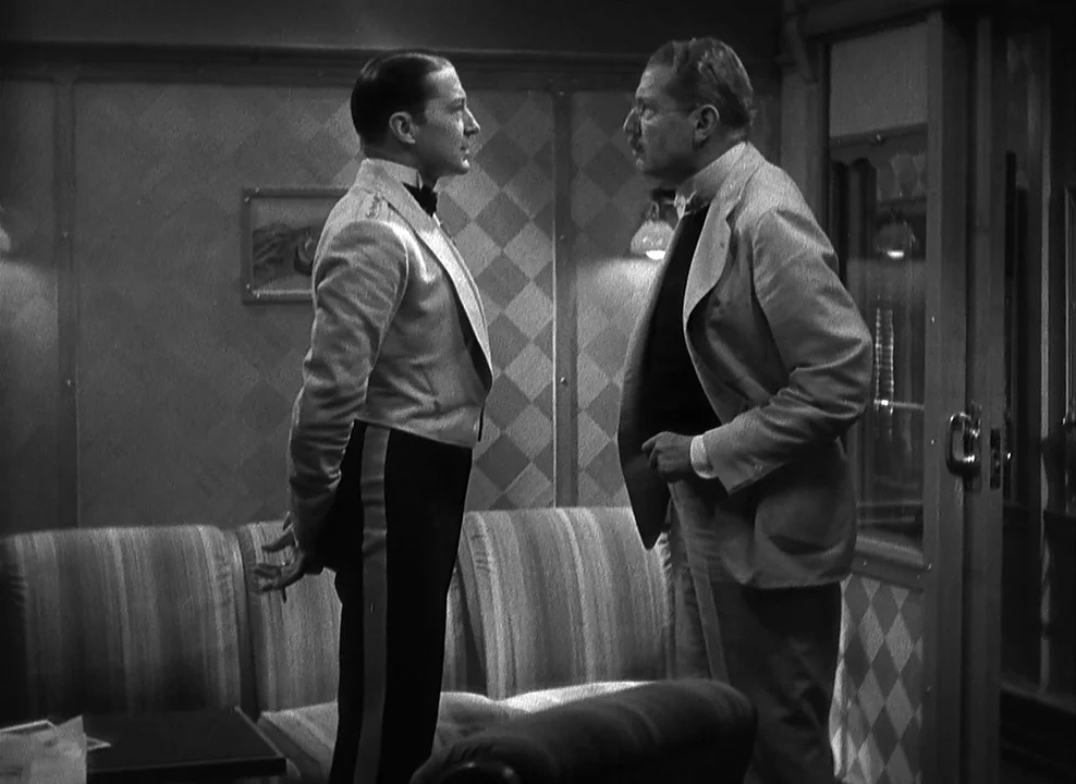 Shanghai Express (1932) movie still: medium — Two men face each other inside a dimly lit railcar compartment with patterned walls. The younger man…