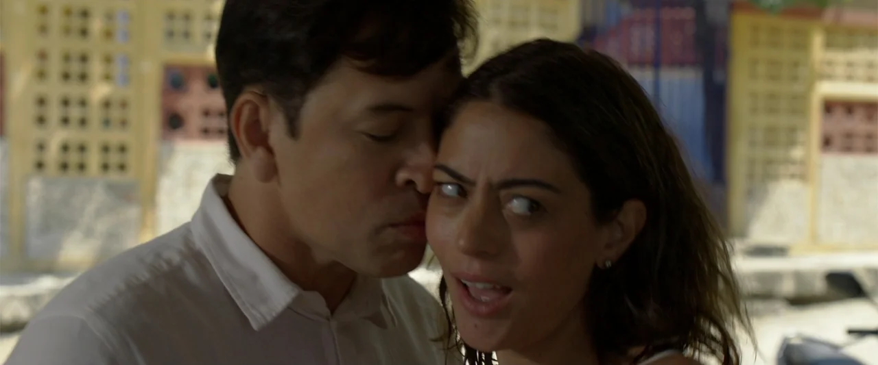 Férias Trocadas (2024) movie still: closeup — An outdoor urban location features a building with yellow walls and grid-pattern vents. A man kisses a…