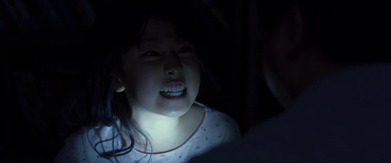 The Wailing (2016) movie still: closeup — A dark, dimly lit room is illuminated only by a small, low-positioned light source. A young girl stares…