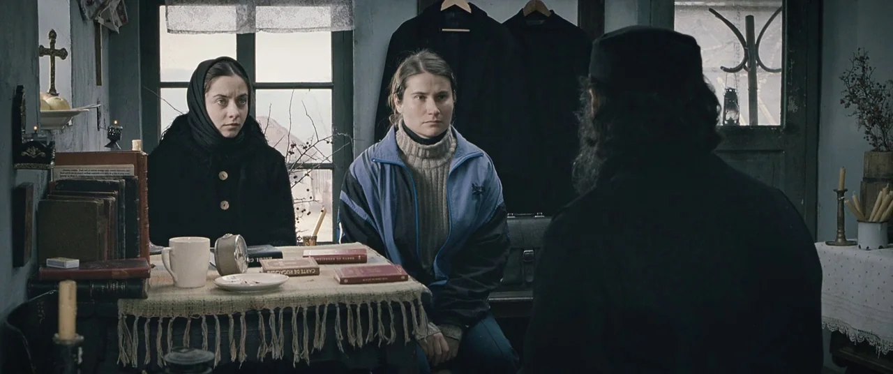 Beyond the Hills (2012) movie still: medium — A spartan room contains a wooden table cluttered with old books and a mug. Two young women sit before…