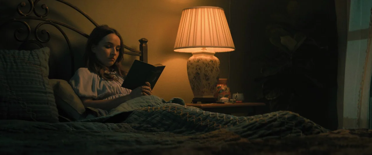 No One Will Save You (2023) movie still: medium — A woman rests in a dimly lit bedroom under a teal quilted blanket. She holds an open book while…