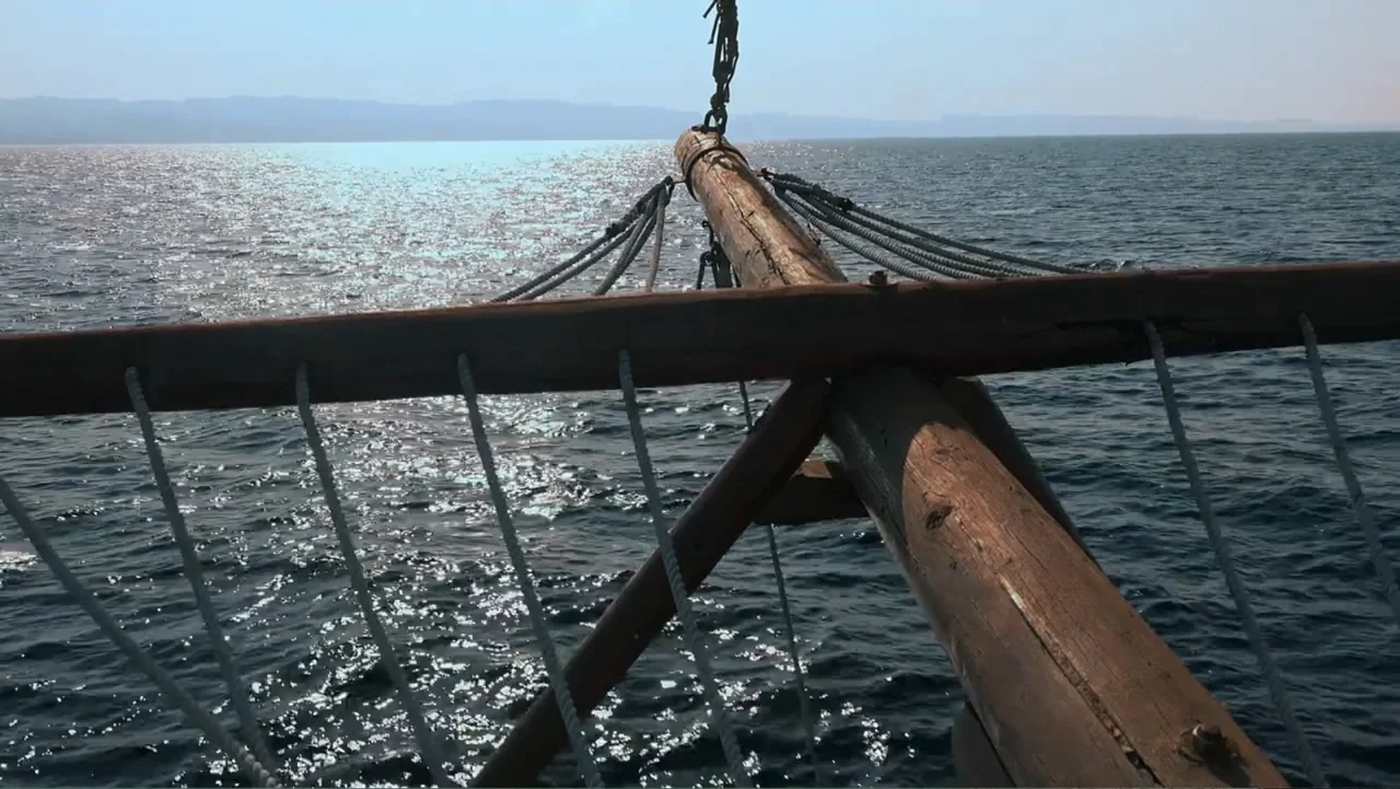 Empire Queen: The Golden Age of Magic (2024) movie still: wide — The bowsprit of a wooden sailing ship extends over a calm sunlit sea. Distant hills…