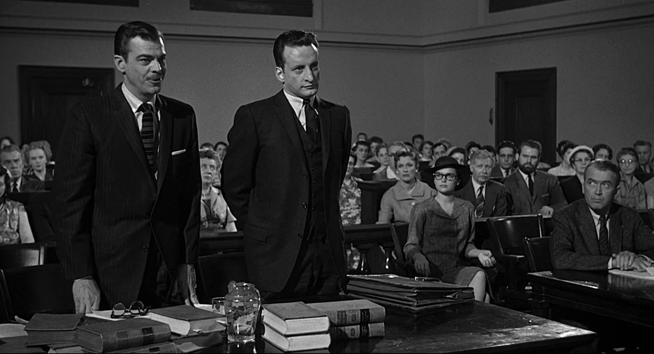 Anatomy of a Murder (1959) movie still: medium — A crowded courtroom features wood paneling and rows of attendees seated in dark benches. Two men…