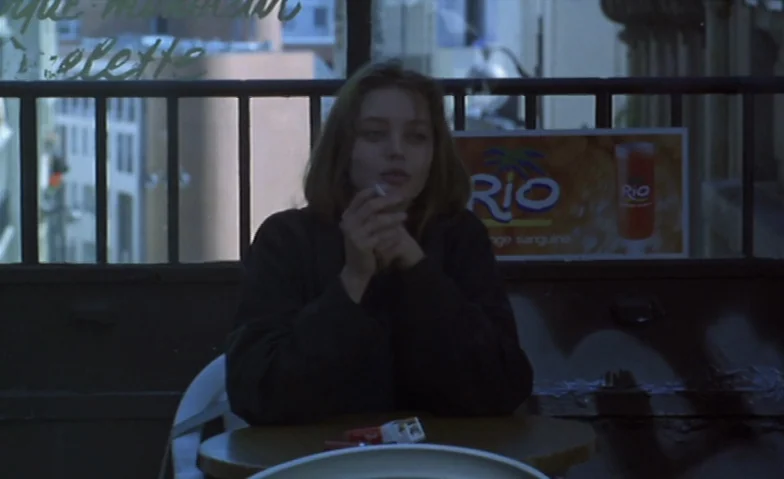 I Can’t Sleep (1994) movie still: medium — An outdoor balcony with a black metal railing and a visible advertisement for juice. A young woman sits…