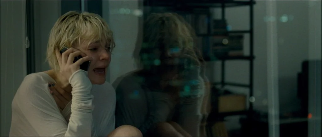 Shame (2011) movie still: medium — A dimly lit modern room at night with city reflections visible on glass. A young blonde woman sits by the window…
