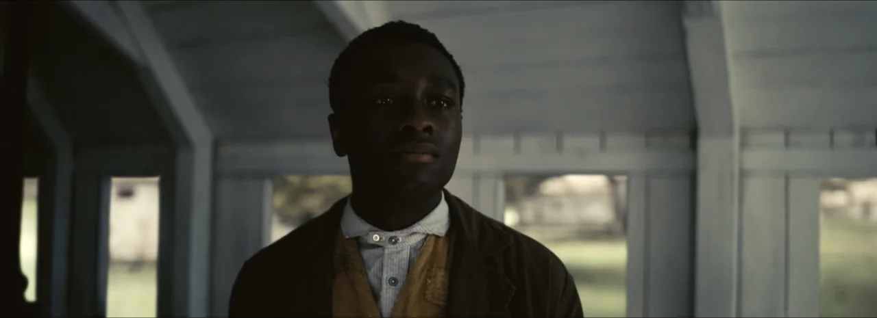 Sinners (2025) movie still: medium — A wooden porch with white framed windows looking out onto a grassy rural area. A young man wearing a vest and…