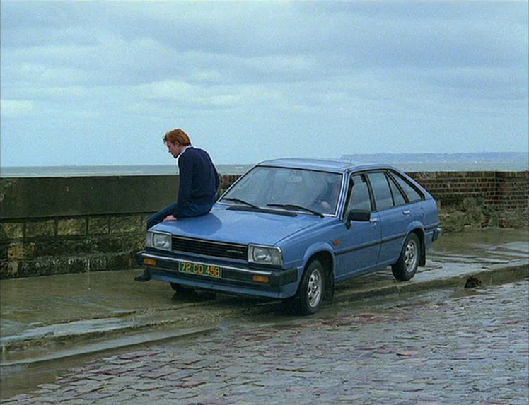 First Name: Carmen (1983) movie still: wide — A man sits on the hood of a blue hatchback beside a stone seawall. An overcast day illuminates the wet…