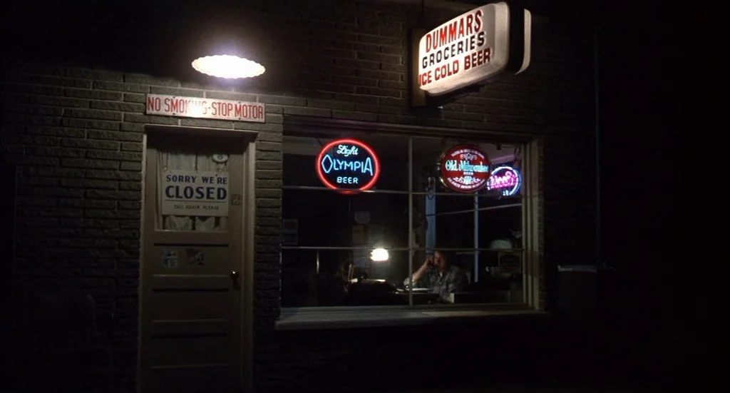 Melvin & Howard (1980) movie still: wide — A small brick grocery store at night with neon signage illuminating the front window. A man sits at a…