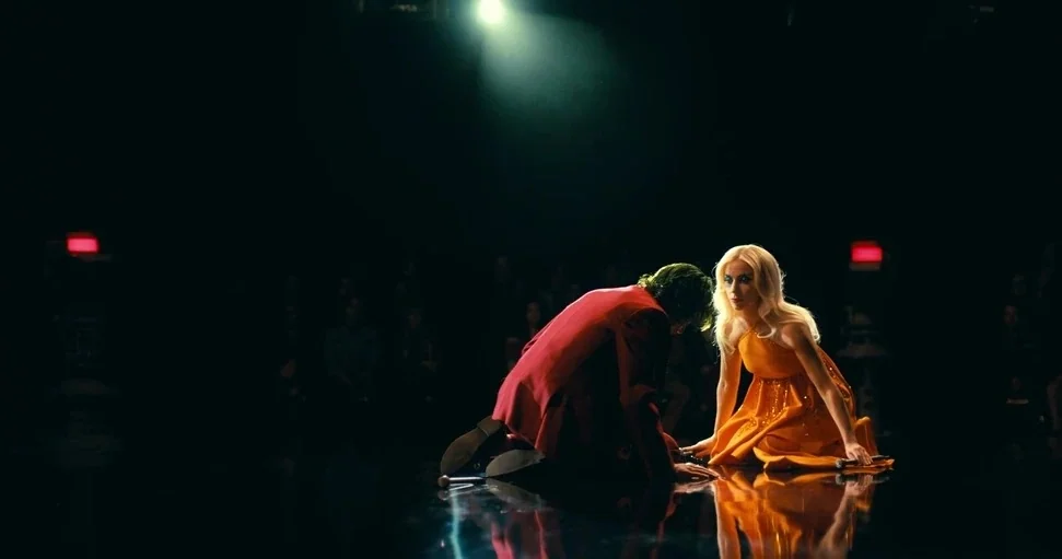 Joker: Folie à Deux (2024) movie still: wide — A dark television studio stage with a reflective black floor under a singular spotlight. A man in a…